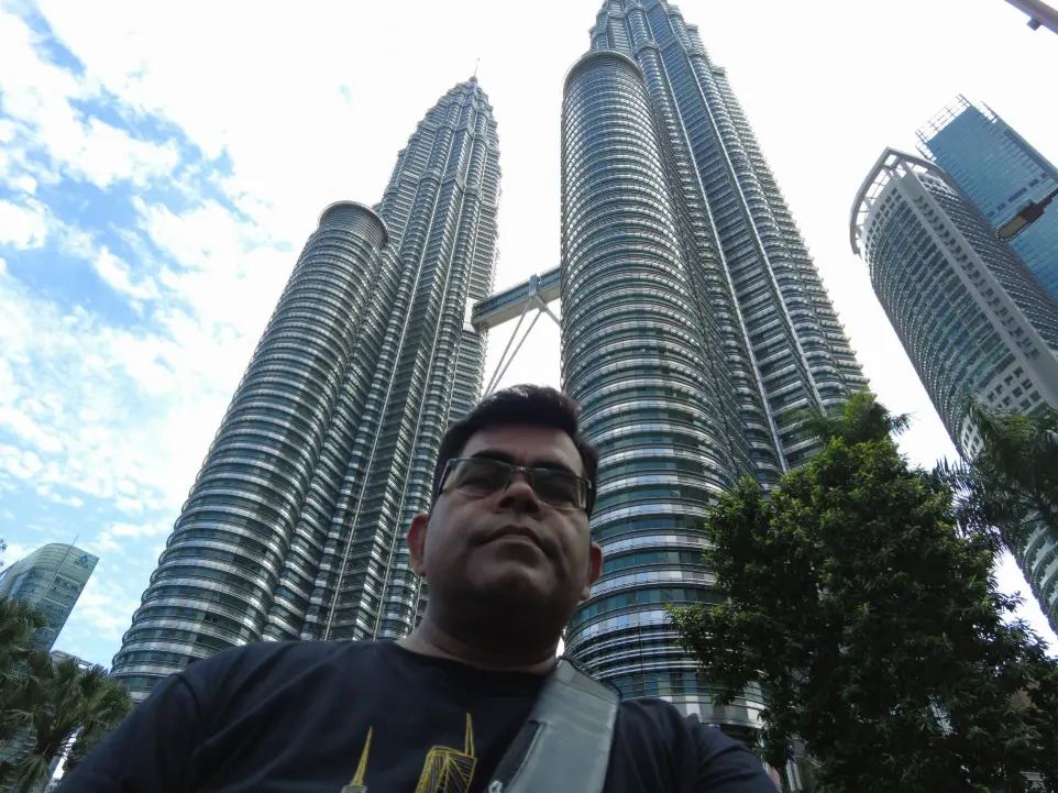 AJ Backpacker is taking a selfie in front of the Petronas Twin Towers in Kuala Lumpur, Malaysia. The angle of the shot captures the impressive height and modern architecture of the towers, with their signature skybridge also visible in the background. AJ is standing in the foreground wearing glasses, a black t-shirt with a golden building design, and a crossbody strap. The photo highlights both the traveler and the iconic landmark, making it a classic tourist moment.
