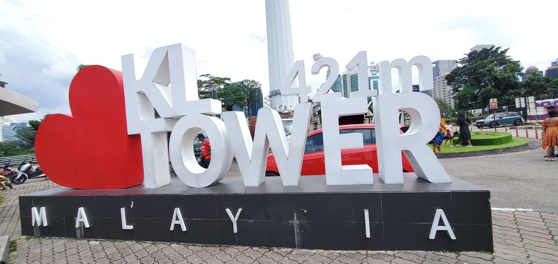 This photo shows the famous I ❤️ KL Tower landmark in Kuala Lumpur, Malaysia. The large 3D letters spell out “KL TOWER 421m” with a big red heart symbolizing love for the city. The background features the KL Tower itself, tall and white, rising above the cityscape. You can also see a few people, vehicles, and buildings around the area, giving the scene a lively and urban vibe.