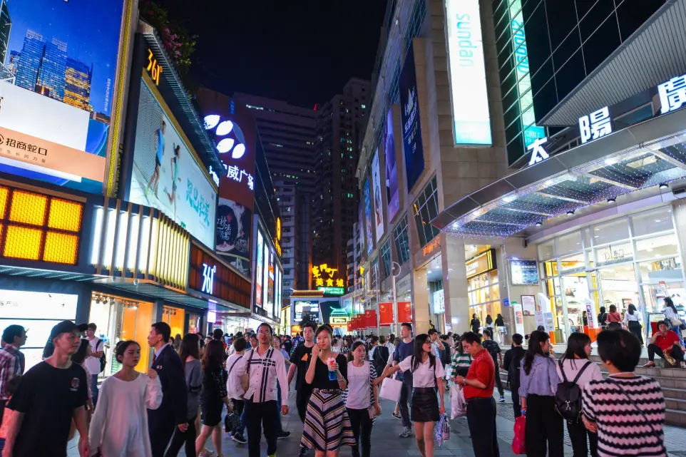 The image shows a bustling city street at night in Shenzhen, China. Bright neon signs, billboards, and LED screens light up the area, creating a lively urban atmosphere. Large shopping malls and stores line both sides of the street, with recognizable brands displayed on glowing signboards.