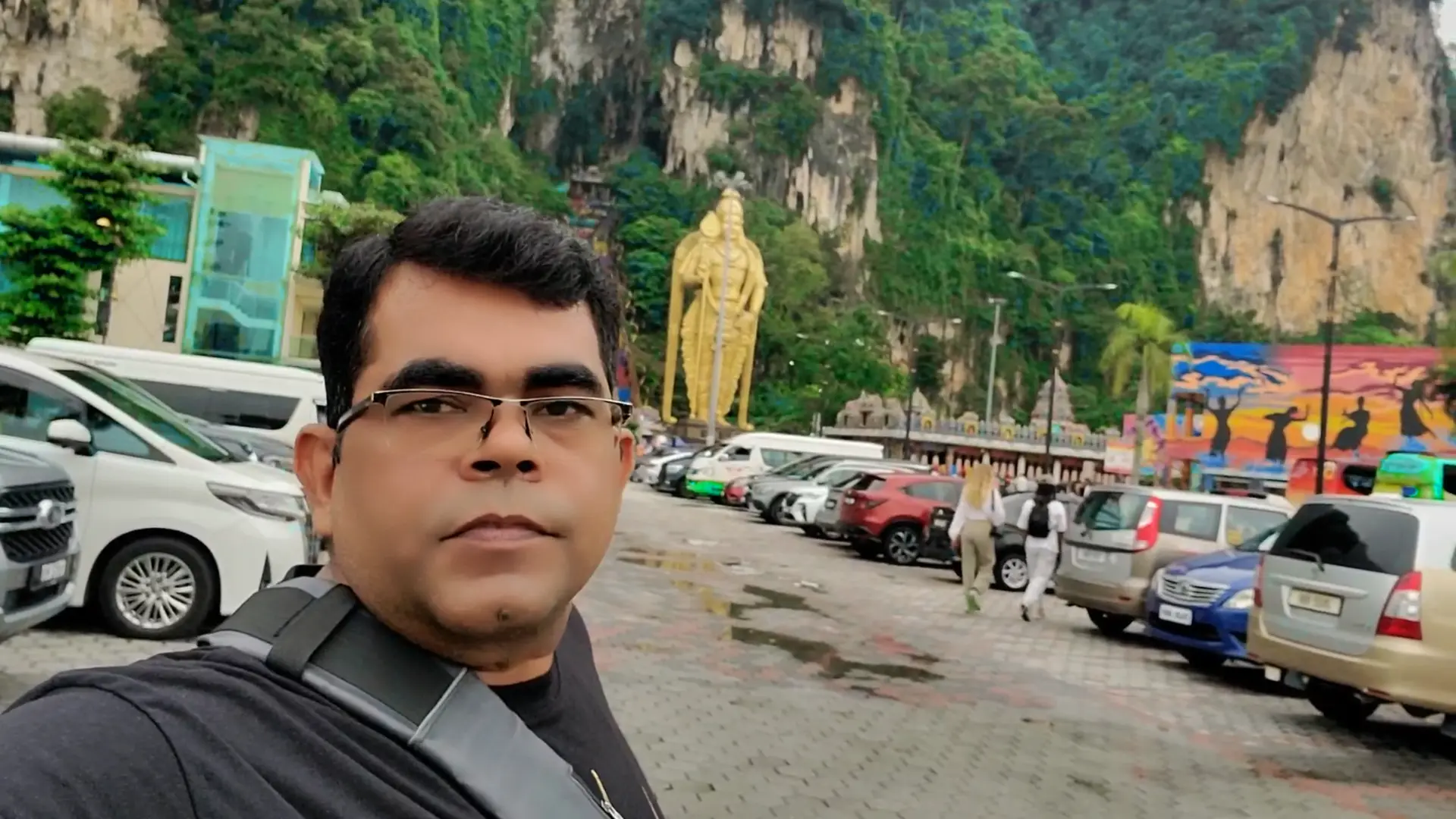 AJ Backpacker is taking a selfie at the Batu Caves in Malaysia. Behind him, the towering golden statue of Lord Murugan stands prominently at the entrance, with the limestone hills and lush greenery forming a dramatic natural backdrop. Several parked cars and visitors can be seen in the area, highlighting the spot’s popularity as a major tourist attraction. The colorful mural on the right adds vibrancy to the scene. AJ is in the foreground wearing glasses, a dark t-shirt, and a crossbody strap, capturing his visit to this iconic cultural and religious landmark.AJ Backpacker is taking a selfie at the Batu Caves in Malaysia. Behind him, the towering golden statue of Lord Murugan stands prominently at the entrance, with the limestone hills and lush greenery forming a dramatic natural backdrop. Several parked cars and visitors can be seen in the area, highlighting the spot’s popularity as a major tourist attraction. The colorful mural on the right adds vibrancy to the scene. AJ is in the foreground wearing glasses, a dark t-shirt, and a crossbody strap, capturing his visit to this iconic cultural and religious landmark.