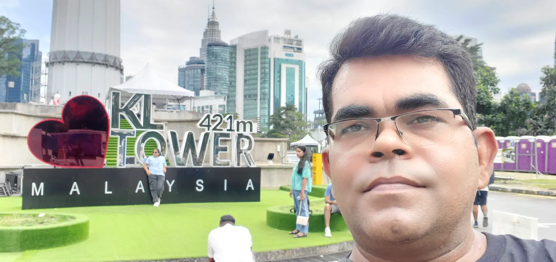 AJ Backpacker is taking a selfie in front of the KL Tower (Kuala Lumpur Tower) signage in Malaysia. The background features the big red heart, the bold &#34;KL TOWER 421m&#34; text, and the word &#34;MALAYSIA&#34; displayed at the base. Behind the sign, the tall tower structure and modern city buildings are visible. Some other tourists can also be seen around the area, adding to the lively atmosphere. AJ is in the foreground, wearing glasses and a black t-shirt, capturing a travel memory at this iconic landmark.